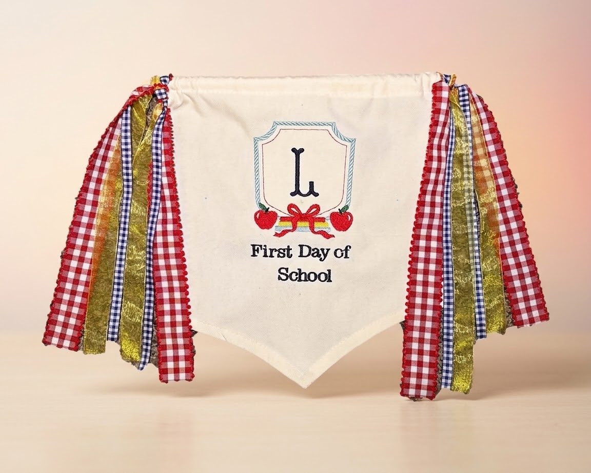 Cream canvas pennant banner reading "First Day of School" with an embroidered monogram and red, gold, and navy ribbon fringe